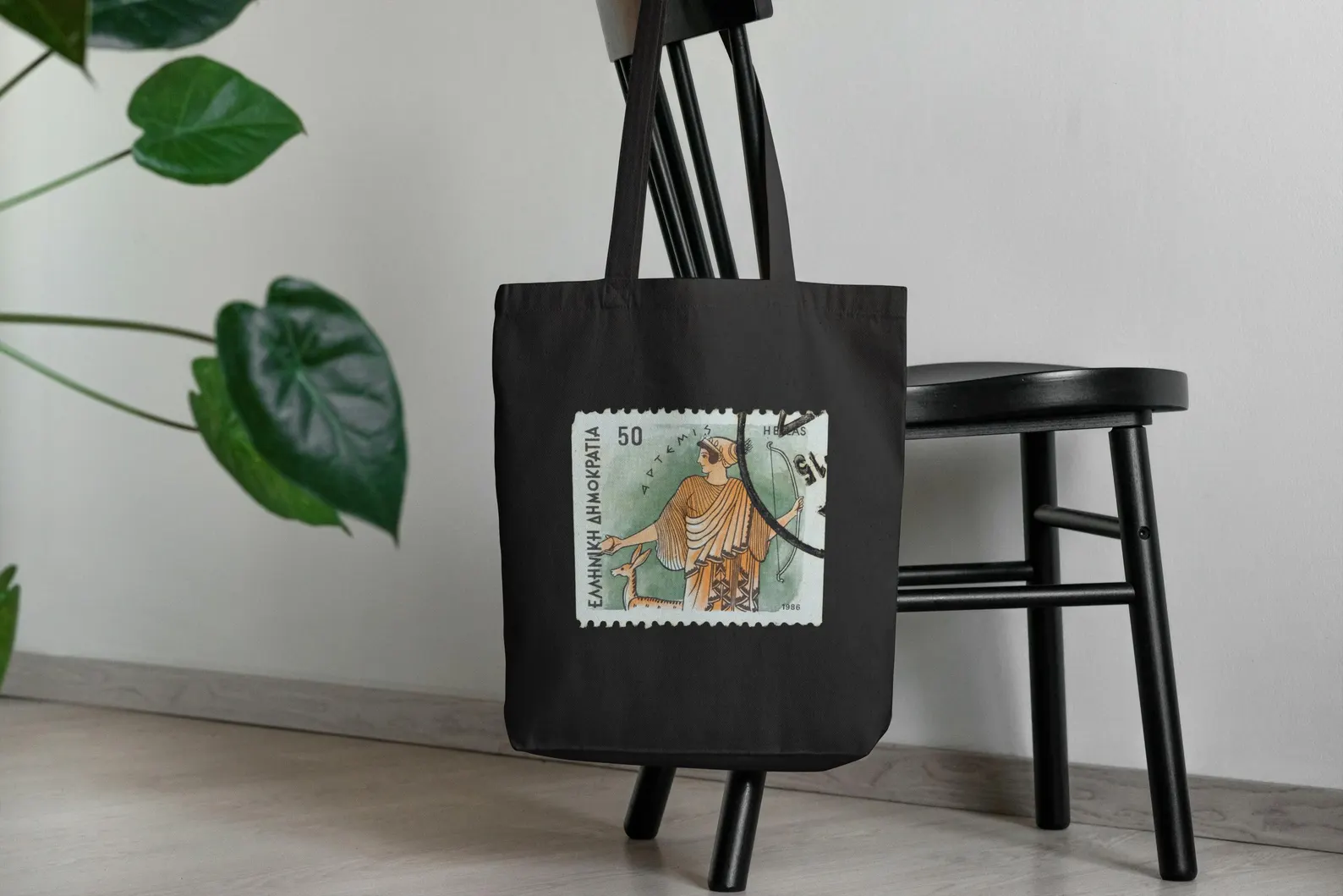 The Artemis goddess tote in black hanging on a chair. A stylish greek mythology home accessory, this goddess of hunting design becomes a conversation starter. Works as a mythology art bag or vintage aesthetic gift for someone who loves Greek history.